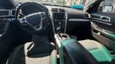 Front interior of a sedan showing the steering wheel, center touchscreen, gear shifter, and black fabric seats with sunlight on the dashboard.