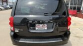 Rear view of a black Dodge Grand Caravan at a dealership, with a Wheel City license plate frame and a silver bumper guard visible under the rear hatch.