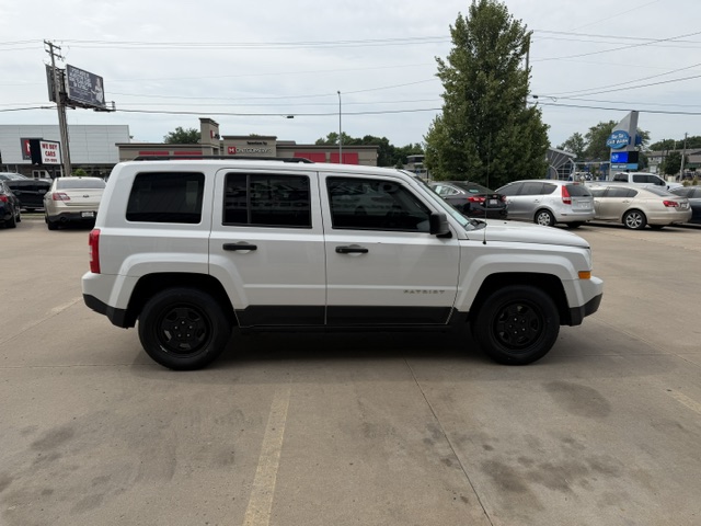 Side view of a white Jeep Patriot SUV parked in a car dealership lot with other vehicles in the background.