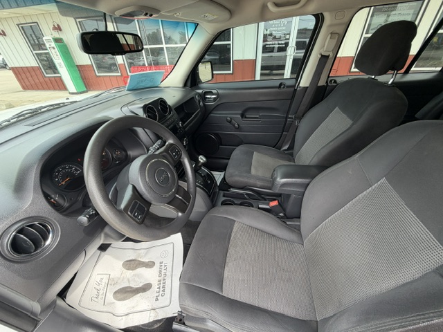 Front interior of a vehicle from the driver's seat, showing steering wheel with a Jeep logo, dashboard, center console, and two gray cloth front seats with a storefront visible outside the windows.