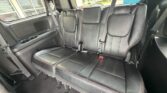 Rear row of a black leather SUV seat with three seating positions, red contrast stitching, and built-in seat belts; cup holders on the left and windows showing a dealership lot outside.