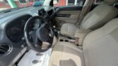 Interior of a beige SUV with the driver’s seat and front passenger seat visible, steering wheel with Jeep logo, center console and cupholders, and outside storefronts through the windows.