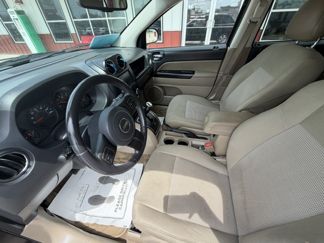 Interior of a beige SUV with the driver’s seat and front passenger seat visible, steering wheel with Jeep logo, center console and cupholders, and outside storefronts through the windows.