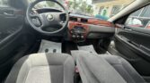 Back-seat view of a Chevrolet sedan interior: gray cloth front seats, wood-trimmed center console, steering wheel, and dashboard; dealership lot visible through the windshield.