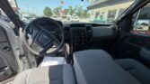 Interior of a gray pickup truck from the driver’s side with the steering wheel, center dashboard, and front bench seat; door open and a dealership lot visible outside.