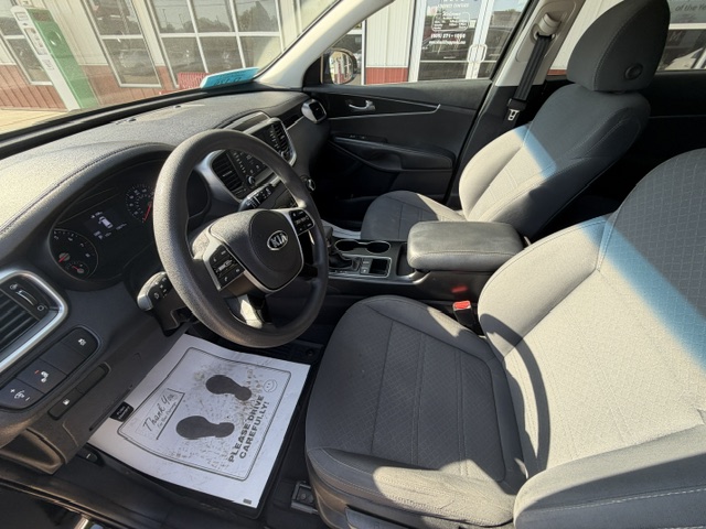Front interior of a gray cloth Kia sedan, showing the steering wheel, dashboard, center console, and front seats bathed in sunlight.