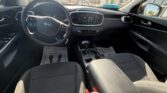 Front seats and dashboard of a Kia car, steering wheel on the left, touchscreen display, gear selector, and floor mats visible in the cabin scene outside a dealership window.