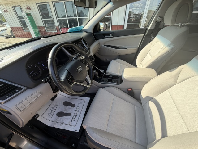 Light gray car interior showing the front seats, steering wheel, dashboard, and center console; driver's door open and paper floor mat visible.
