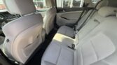 Interior of a light-gray sedan viewed from the rear seat, showing a three-seat rear bench, seat belts, and front seats with storage pockets; sunlight on the upholstery.