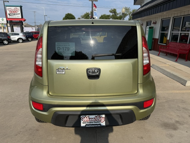 Rear view of an olive-green Kia Soul in a dealership lot, with a Wheel City license plate frame and a back window sticker visible.
