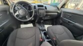 Front interior of a Kia: steering wheel, center console with display, gear shifter, and two dark cloth seats; view of a parking lot through the windows.