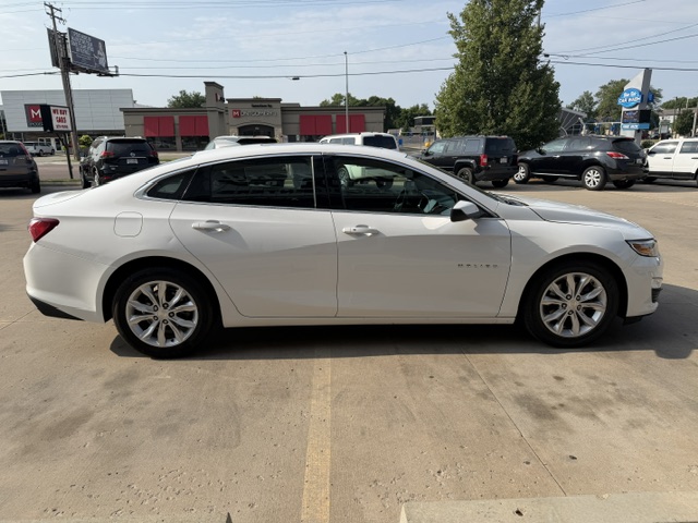 Side view of a white four-door sedan parked in a lot, with other vehicles and a strip mall in the background.