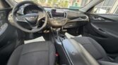 Front interior of a Chevrolet sedan showing steering wheel, center console with touchscreen, gear shifter, and black cloth seats.