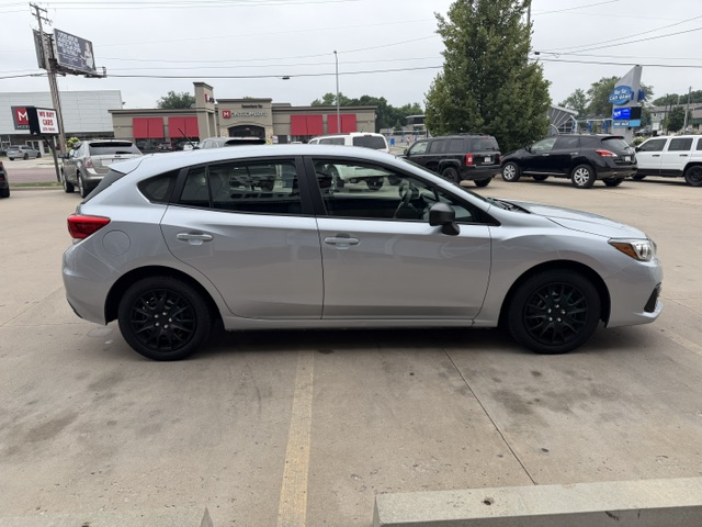 Side view of a silver hatchback car with black wheels parked in a parking lot, with other vehicles and storefronts in the background.