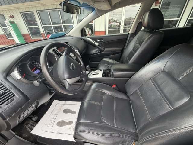 Front interior of a black leather SUV showing the dashboard, steering wheel, center console, gear shifter, and both front seats; a floor mat with footprints is visible on the driver’s side.