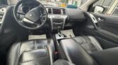 Interior view of a black sedan from the driver’s seat: leather front seats, steering wheel with Nissan logo, center console with automatic gear shifter, and dashboard; dealership lot visible through the windows.