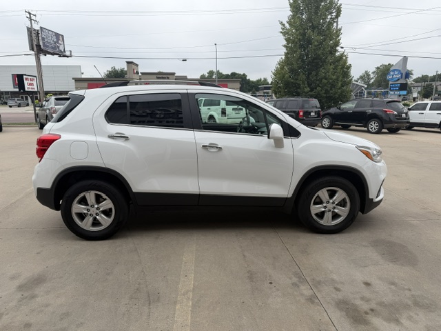 Side view of a white compact SUV parked in a lot, with black lower trim and silver alloy wheels, and other vehicles and shops in the background.