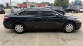 Black four-door sedan parked in a dealership lot with other cars and storefronts in the background, side view. (Informative)