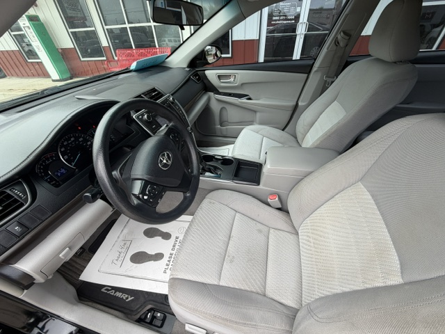 Interior view of a Toyota Camry from the driver’s side, showing the steering wheel, dashboard, center console, and light gray cloth seats. A paper floor mat with footprints rests on the driver’s side.