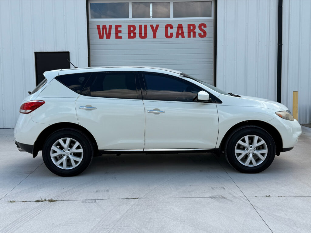 White SUV parked in front of a building with a large 'WE BUY CARS' sign on a gray garage door behind it.