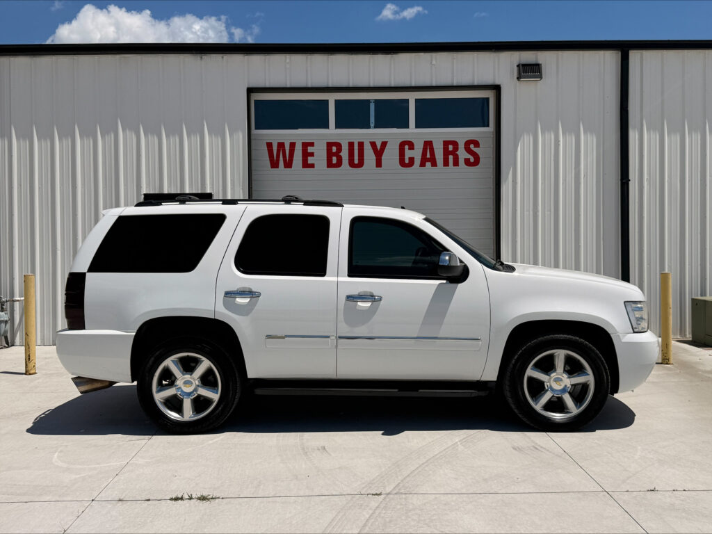 White SUV parked in front of a building with a large garage door reading 'WE BUY CARS' in red letters.
