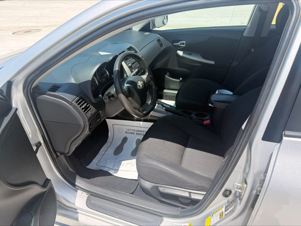 Interior of a car from the driver's side showing the steering wheel, dashboard, center console, and papers on the floor mat near the pedals