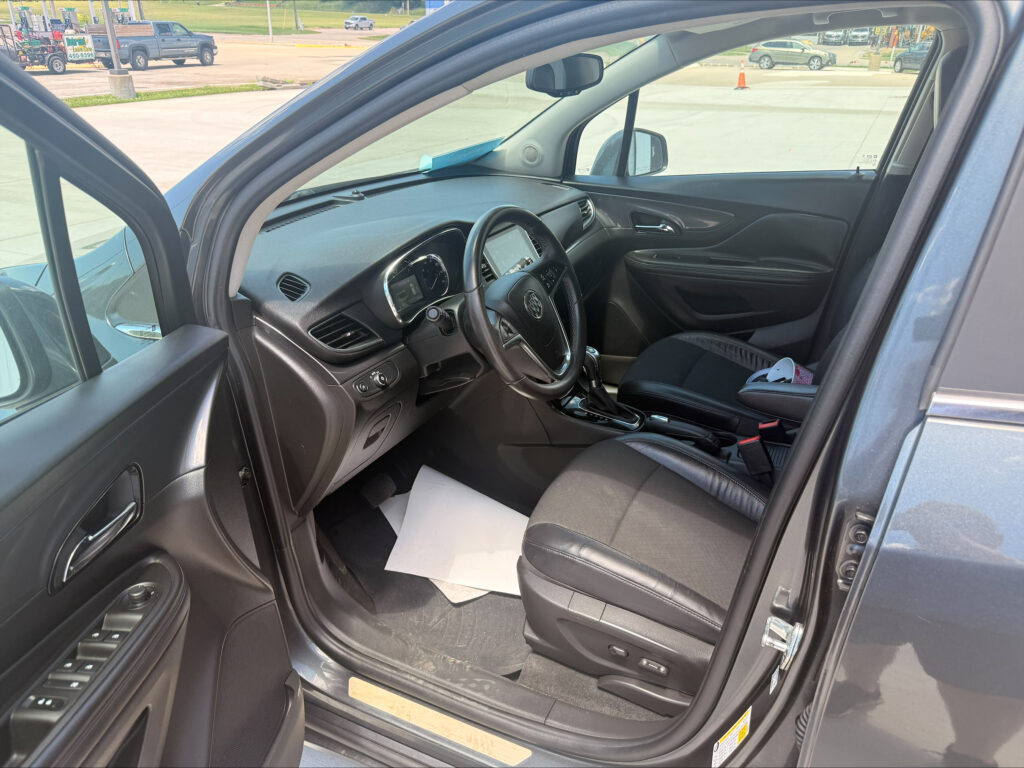 Open-car interior: driver’s side view with steering wheel, dashboard, and center console visible; outside parking lot scene through windows.