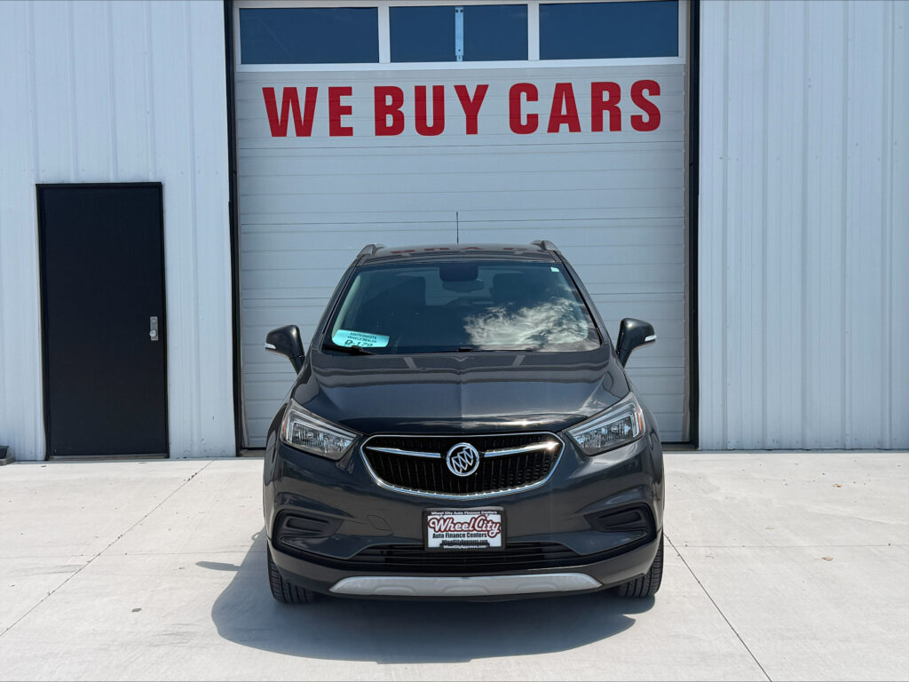 Gray Buick SUV parked in front of a white garage door with a large red 'WE BUY CARS' sign above it