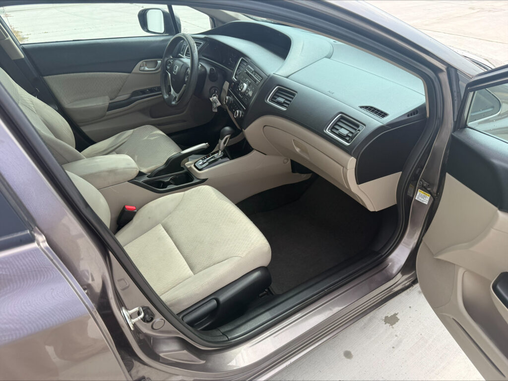 Interior view of a Honda sedan from the open front passenger door, showing beige cloth seats, steering wheel, center console, and dashboard.