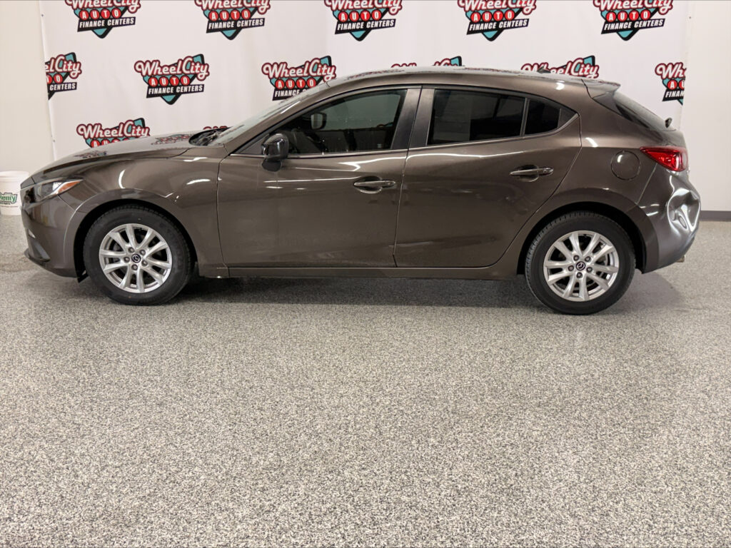 Side view of a brown sedan parked indoors with a Wheel City Auto backdrop behind it, showing four doors and alloy wheels.