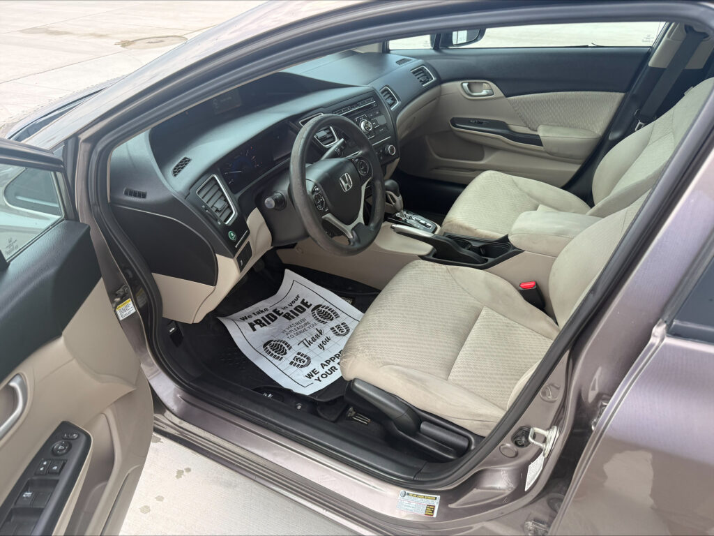 Interior of a beige car with the driver’s seat, steering wheel, and center console visible through the open front door; floor mat on the driver side.