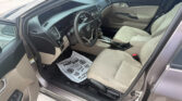 Interior of a beige car with the driver’s seat, steering wheel, and center console visible through the open front door; floor mat on the driver side.