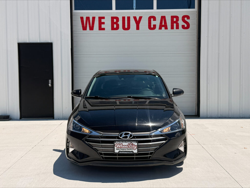 Front view of a black Hyundai sedan parked in front of a white building with a large 'WE BUY CARS' sign above the bay door