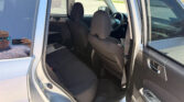 Interior of a silver SUV with the rear door open, showing black fabric seats and the steering wheel in front; a person’s reflection is visible in the window as they take a photo in daylight.