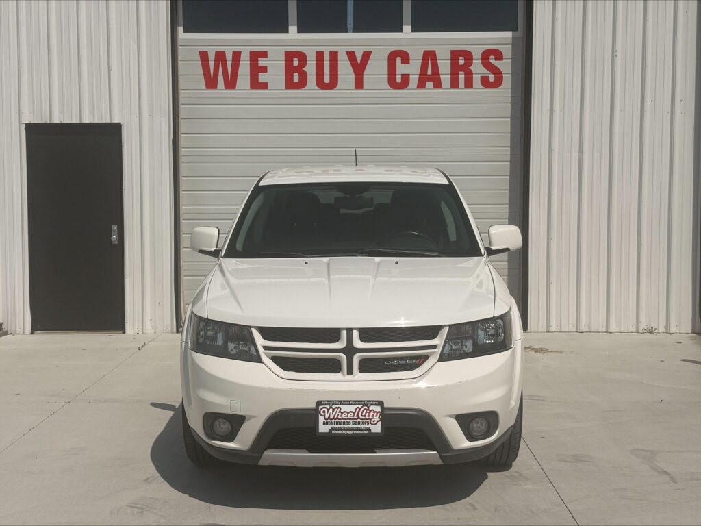 White Dodge SUV head-on in front of a garage door that says 'WE BUY CARS' in red letters.