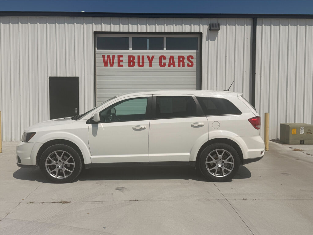 White SUV parked in front of a metal-sided building with a large garage door reading 'WE BUY CARS' under a row of windows.
