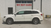 White SUV parked in front of a metal-sided building with a large garage door reading 'WE BUY CARS' under a row of windows.