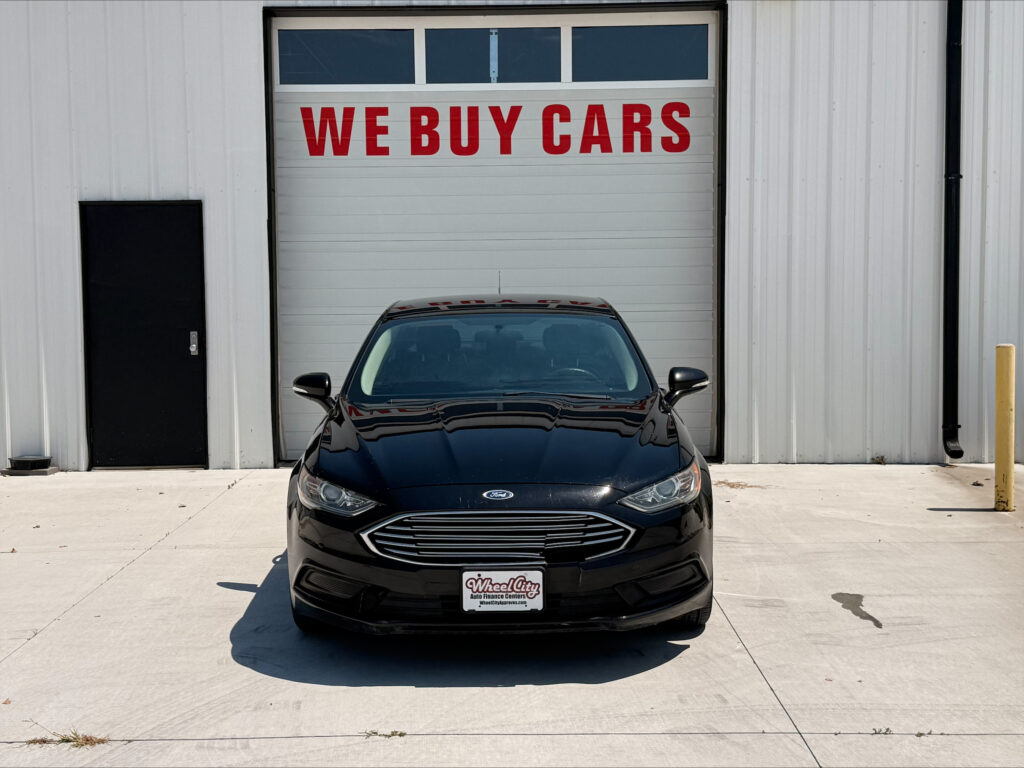 Black Ford sedan facing the camera, parked in front of a garage with a large red 'WE BUY CARS' sign on the door.