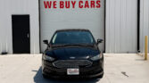 Black Ford sedan facing the camera, parked in front of a garage with a large red 'WE BUY CARS' sign on the door.