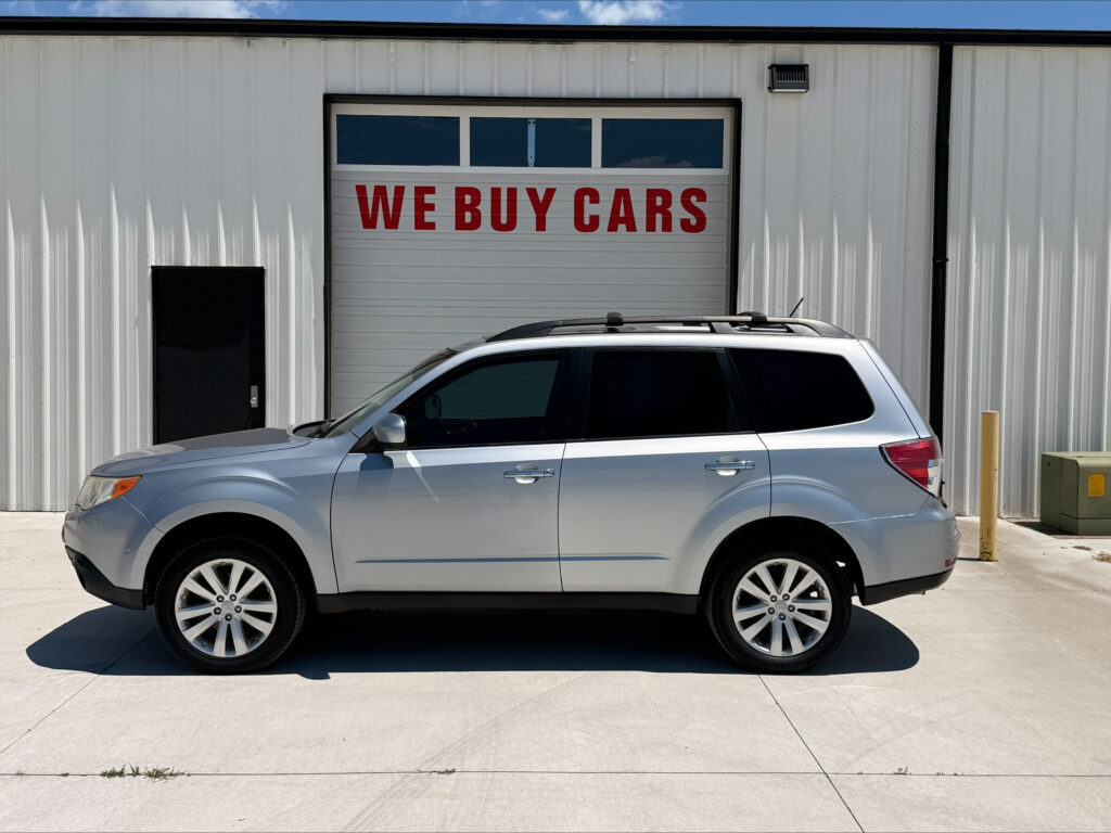 Silver SUV parked in front of a light-colored industrial building with a large garage door reading 'WE BUY CARS'.