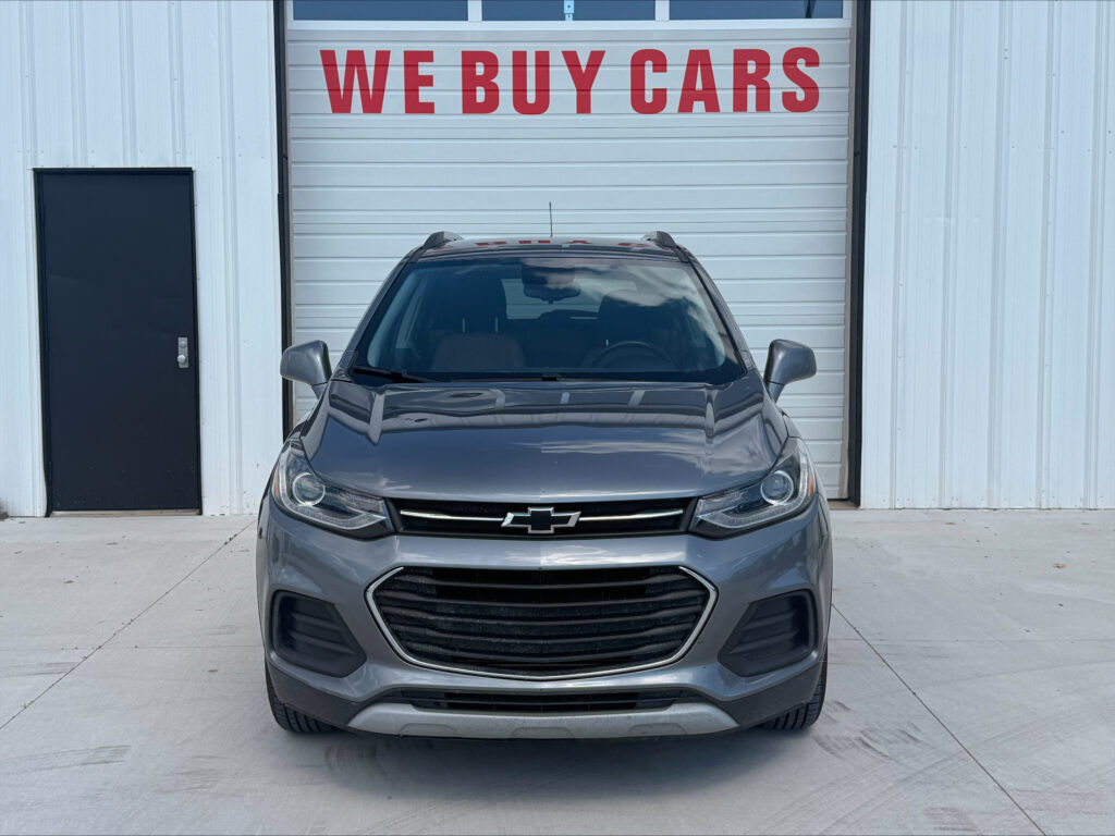 Front view of a gray Chevrolet SUV parked in front of a white building with a large red 'WE BUY CARS' sign on the door behind it.