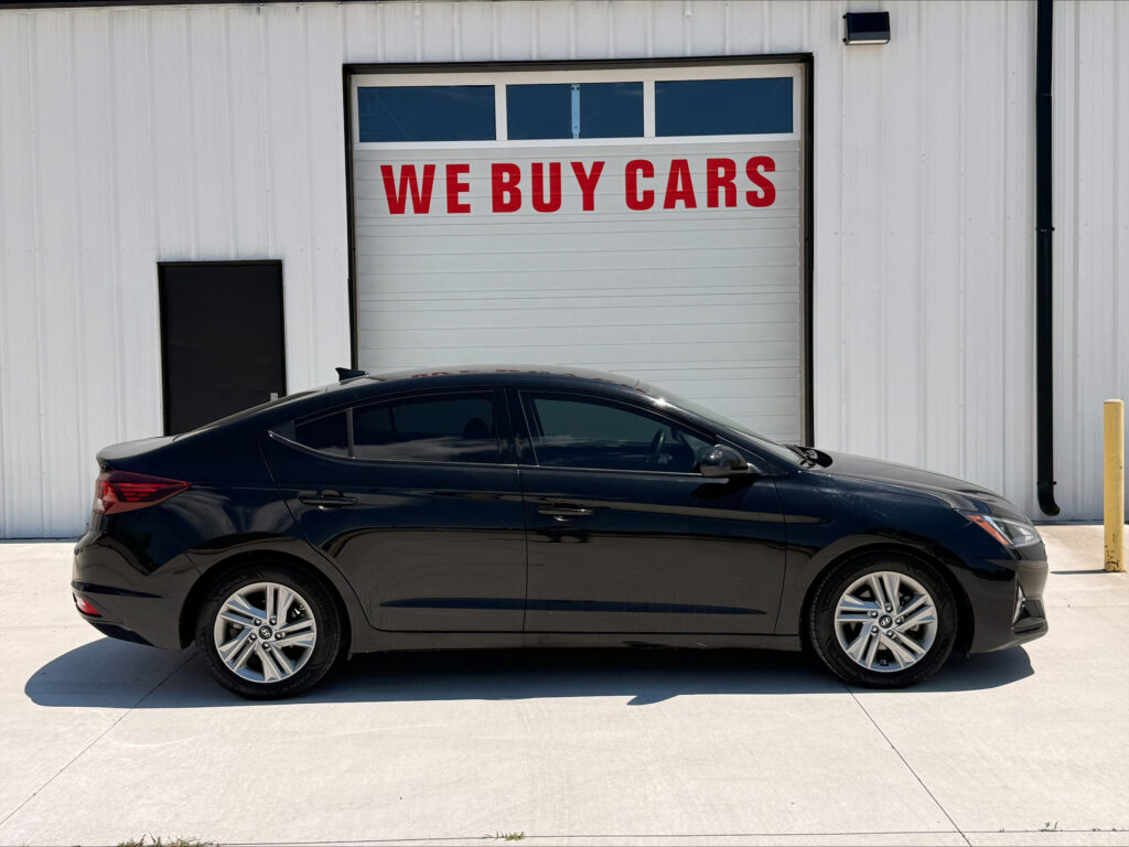 Black sedan parked in front of a white industrial building with a large garage door reading 'WE BUY CARS' in red lettering.