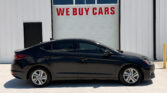 Black sedan parked in front of a white industrial building with a large garage door reading 'WE BUY CARS' in red lettering.