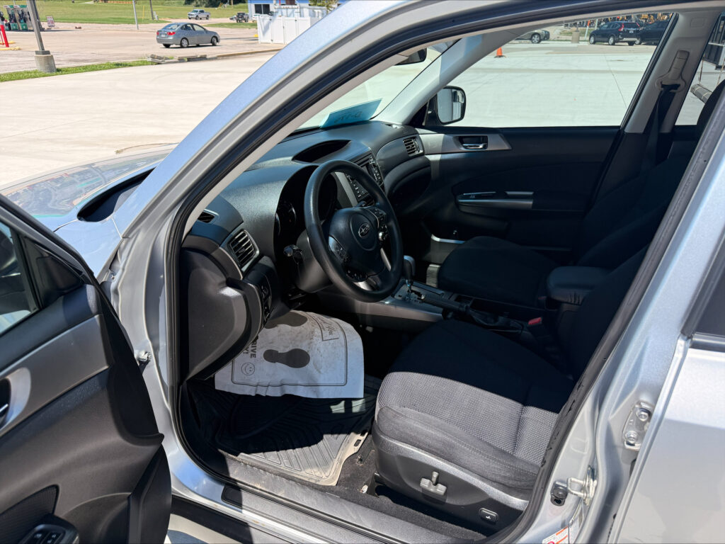 Interior of a silver SUV with the driver's-side door open, showing the steering wheel, dashboard, and center console; parking lot outside.