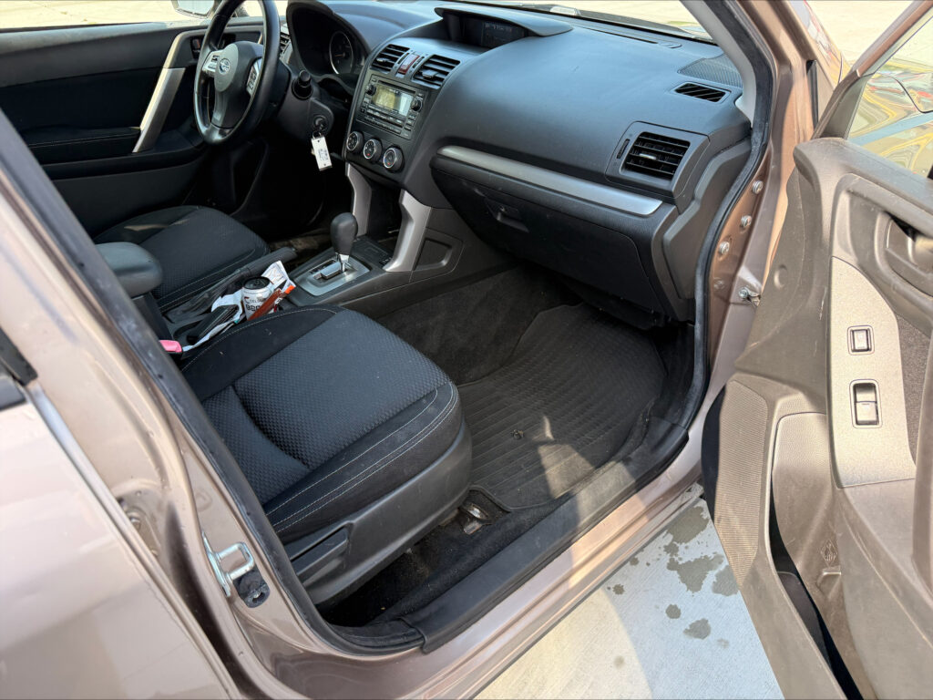 Interior of a car showing the driver's seat, steering wheel, center console, and a can and papers on the floor beside the gear shifter.