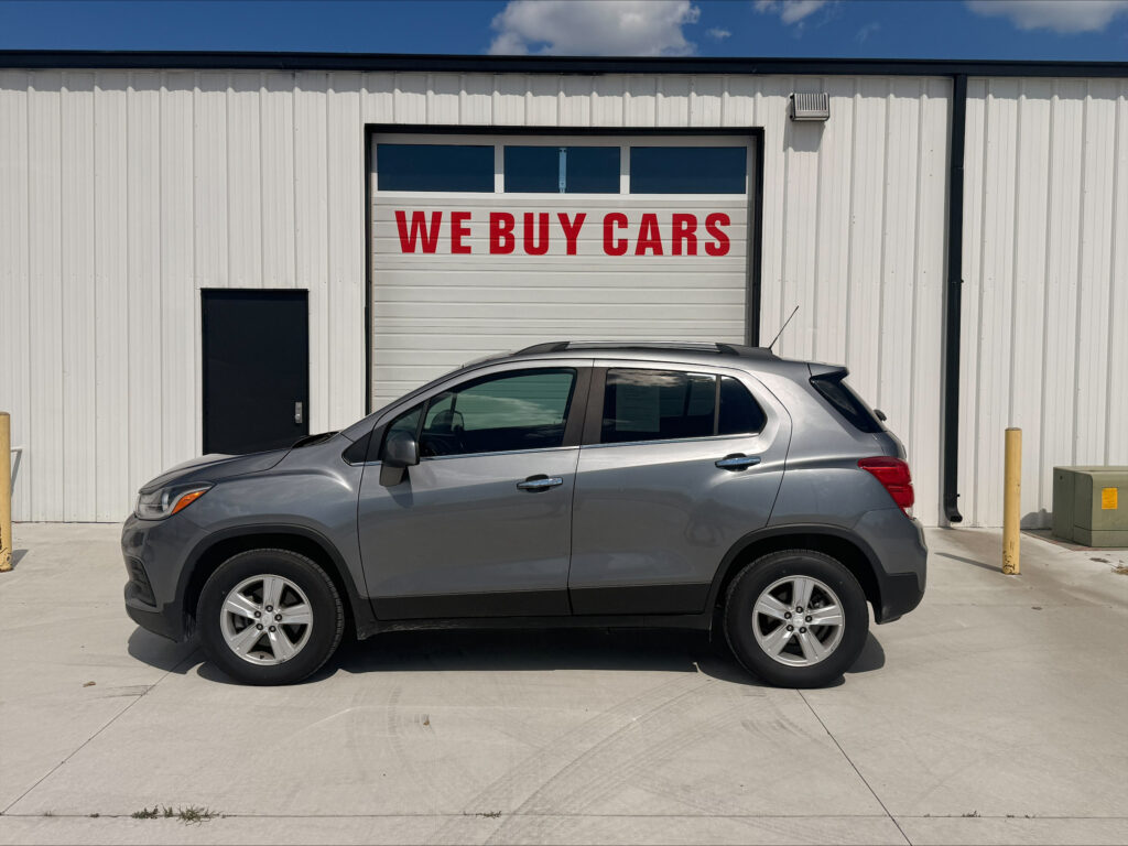 Silver SUV parked in front of a white metal building with a large garage door displaying 'WE BUY CARS' in red letters.