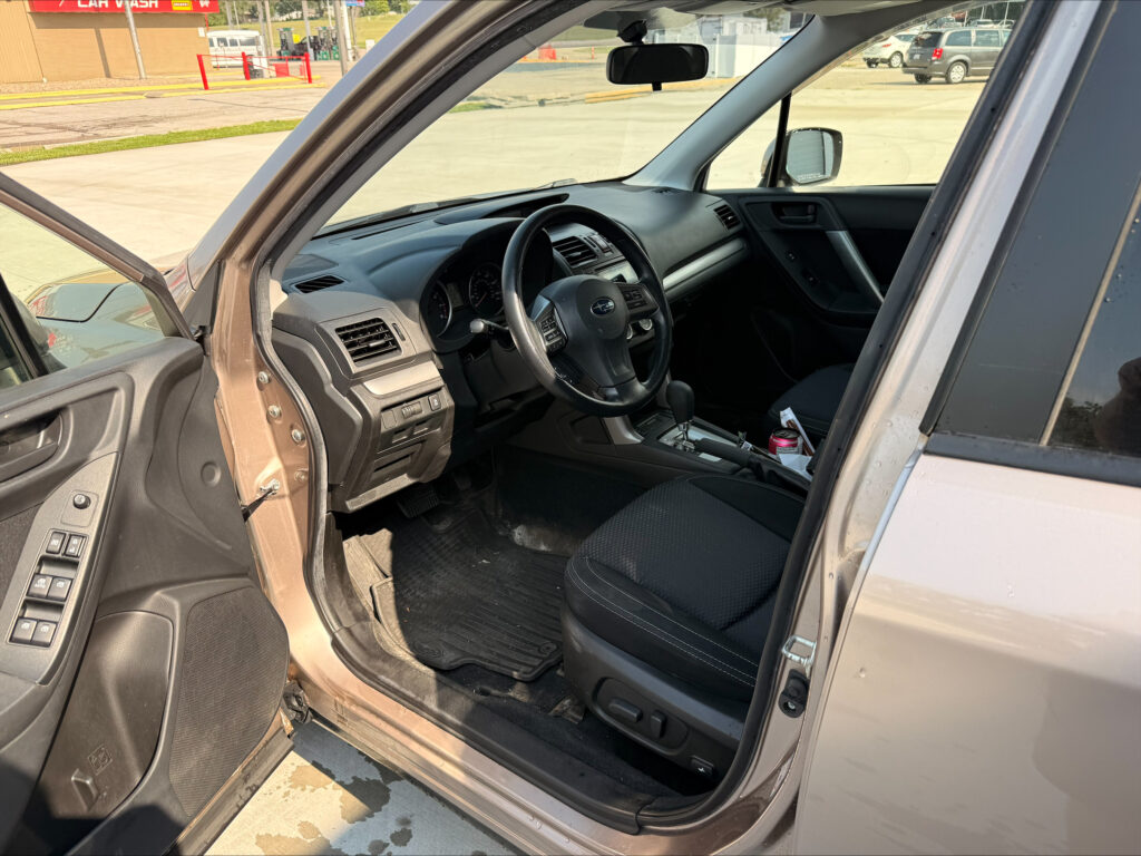 Interior of a car with the driver's door open, showing the steering wheel, dashboard, and center console; gas station lot visible outside.