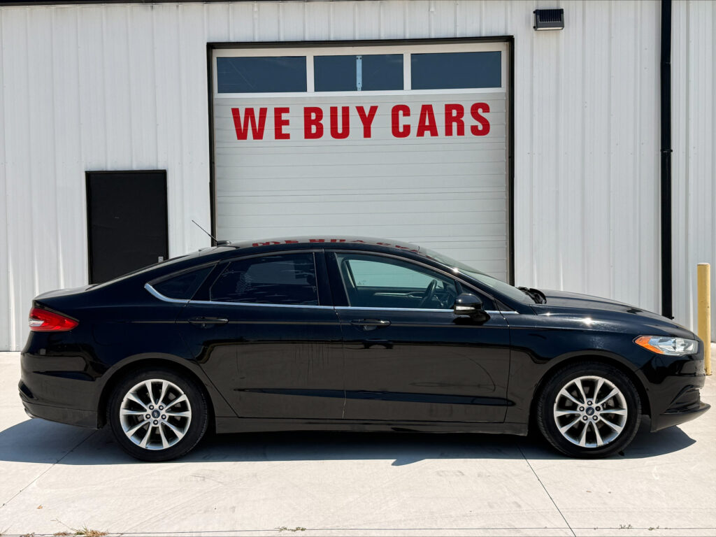 Black sedan parked in front of a white industrial building with a large garage door reading 'WE BUY CARS' in red letters above the car window line.