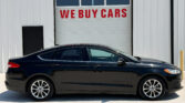Black sedan parked in front of a white industrial building with a large garage door reading 'WE BUY CARS' in red letters above the car window line.