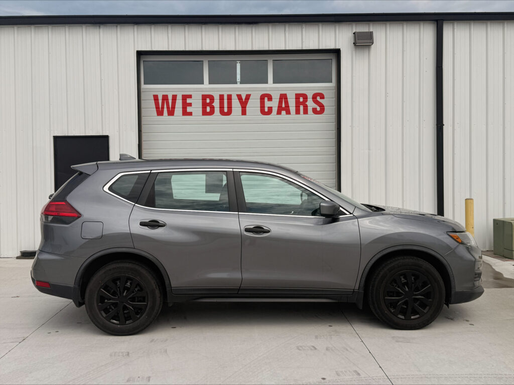 Gray SUV parked in front of a white metal warehouse with a garage door that reads 'WE BUY CARS'.